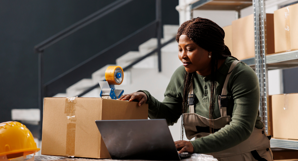 A woman is in her workshop wearing an apron, with boxes and working on her laptop